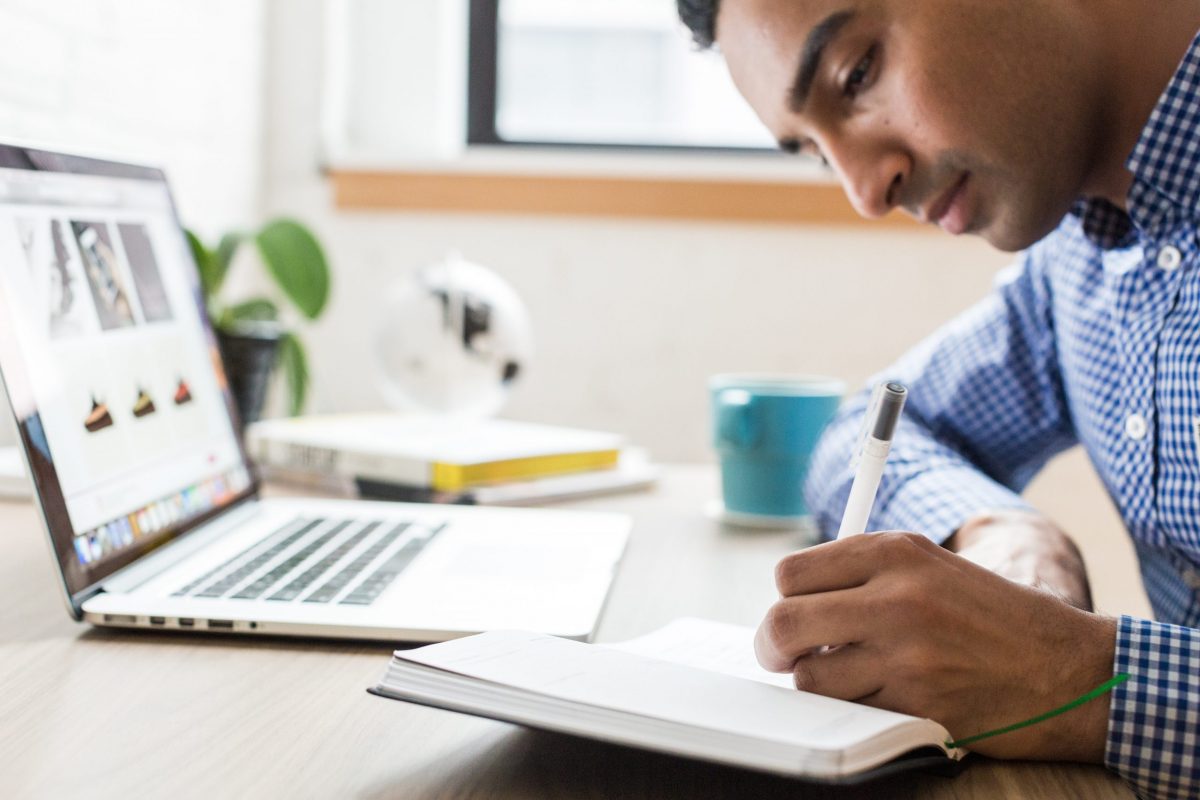 Man writing in a journal at his desk with his laptop open.